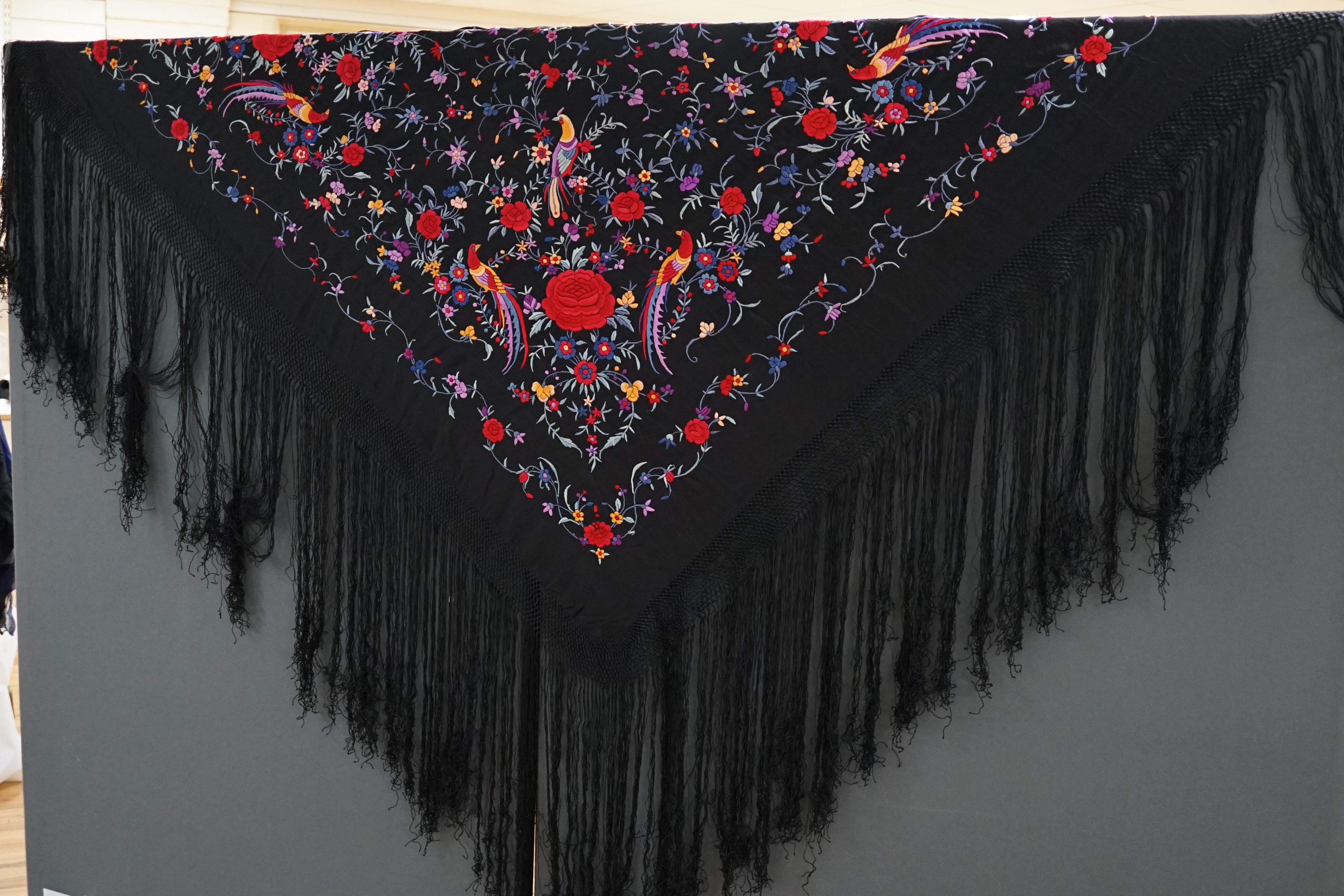 An early 20th century Chinese black silk fringed shawl with polychrome silk embroidery, embroidered with meandering floral design, the corners embroidered with three brightly coloured birds of paradise, large red roses a